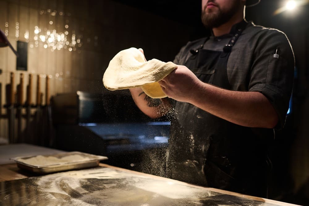 A person in an apron stretches pizza dough by hand in a dimly lit kitchen, with flour dusting the surface and a tray of dough nearby.